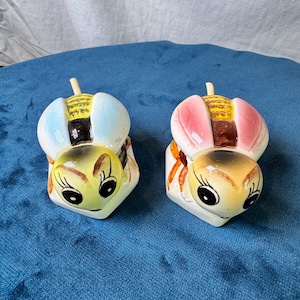 May include: Two ceramic bee-shaped shakers featuring yellow heads with black painted eyes and antennae. One bee has light blue wings, while the other has pink wings. Both have yellow and black striped bodies and rest on white hexagonal bases.