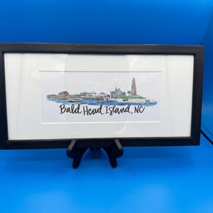 May include: A framed panoramic watercolor print featuring a coastal landscape of Bald Head Island, NC, with a lighthouse, buildings, and a boat on the water. The artwork is set within a white mat and a black rectangular frame.