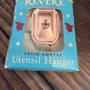 May include: A vintage Revere solid copper utensil hanger packaged in a light blue cardboard box. The box features the text REVERE and SOLID COPPER UTENSIL HANGER with red silhouettes of a head and white stars. The copper hanger is visible.