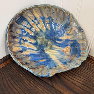 May include: A handcrafted ceramic serving dish with a seashell design. The dish features a blend of blue, tan, and cream colors, with a glossy finish. The rim has a light blue border. The dish is approximately 12 inches in diameter.