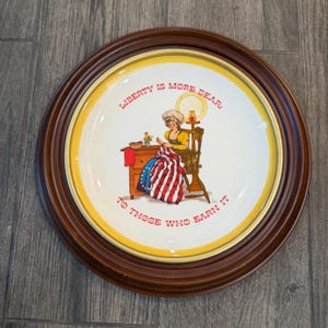 May include: A round ceramic decorative plate with a dark brown wooden frame. The center features a cream-colored surface with a yellow border, depicting a person sewing an American flag. The text reads: LIBERTY IS MORE DEAR TO THOSE WHO EARN IT.