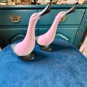 May include: A pair of ceramic goose figurines featuring a smooth, matte finish. Each goose has a soft pink body with dark grey accents on the wings, tail, and beak. The birds are depicted with long, slender necks extended upward in a graceful pose.
