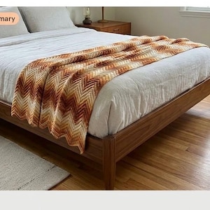 May include: A crochet throw blanket featuring a chevron pattern in gradient shades of brown, orange, and cream. The blanket is draped across a white bedspread on a wooden bed frame, showcasing a textured, zigzag stitch design throughout the piece.