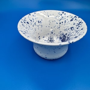 May include: A white ceramic bowl with a wide, flared rim and a central conical spike. The bowl features a speckled pattern of dark blue spots across its entire interior and exterior surface, creating a distinct contrast against the white base.
