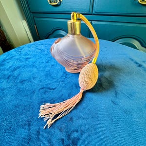 May include: A vintage pink perfume bottle with a gold-colored metal top and a matching atomizer. The bottle has a frosted finish and a decorative design. The atomizer bulb and tassel are a light peach color. The bottle is on a blue velvet surface.