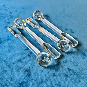 May include: Four clear glass spoons with rounded bowls and rectangular handles are arranged diagonally on a blue surface. The spoons are transparent, allowing the blue background to be visible through them. The spoons appear to be for serving or decorative purposes.