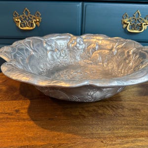 May include: A silver-colored, decorative bowl with a scalloped edge and embossed floral and animal designs. The bowl is round and sits on a wooden table. The background includes a dark blue cabinet with gold hardware.
