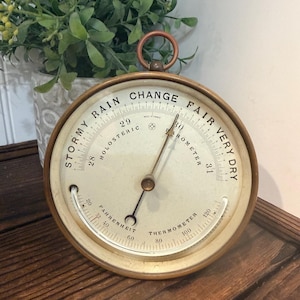 May include: A round brass-toned holosteric barometer and thermometer with a cream-colored face. The dial features black text reading STORMY, RAIN, CHANGE, FAIR, VERY DRY, and FAHRENHEIT THERMOMETER. It includes a top hanging ring and two needles.