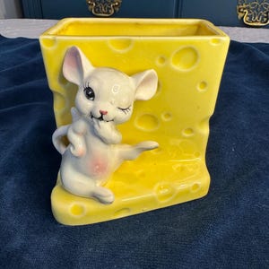 May include: A yellow ceramic planter shaped like a block of cheese, with a winking white mouse figurine attached. The mouse has pink accents on its ears and belly. The planter is rectangular with rounded edges.