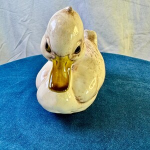 May include: A ceramic duck figurine featuring a glossy off-white body with subtle brown mottled detailing and a smooth, dark yellow-brown beak. The duck is depicted in a seated position with its head tilted slightly forward and dark eyes.
