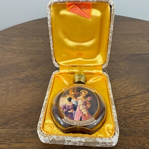 May include: A vintage perfume bottle with a decorative label featuring a painting of women in period clothing. The bottle is housed in a gold satin-lined case with a patterned exterior. The label reads "Jean Desprez".