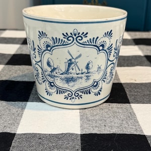 May include: A white ceramic planter with a blue and white windmill design. The planter has a cylindrical shape with a slightly flared rim. The design features a detailed landscape scene with windmills, houses, and trees. The planter sits on a black and white checkered surface.