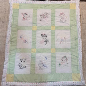 May include: A handmade baby quilt featuring nine embroidered squares with images of children and pets. The quilt has a light green center, yellow corner squares, and a white border with pink polka dots. The embroidery is in shades of red, blue, and black.