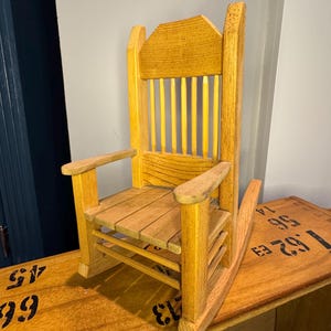 May include: A small, wooden rocking chair with a natural finish. The chair features a slatted seat and back, armrests, and curved rockers. The wood grain is visible, and the chair appears to be handcrafted. A classic, miniature furniture piece.