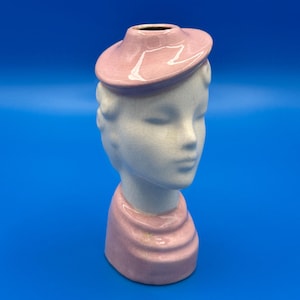 May include: A ceramic head vase featuring a white face with a fine crackle glaze finish. The figure wears a pink hat that serves as the vase opening and a matching pink collar at the base. The sculpture depicts a stylized head and neck.