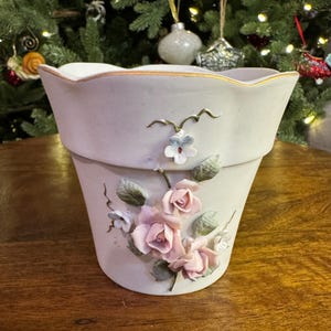 May include: A light-colored ceramic flower pot with a scalloped rim and gold trim. The pot is decorated with raised pink roses, small blue flowers, and green leaves. The pot is sitting on a wooden surface.