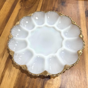 May include: A white, scalloped deviled egg serving dish with a gold-colored trim. The dish has 12 individual egg compartments surrounding a central serving area. The dish is made of glass and sits on a wooden surface.