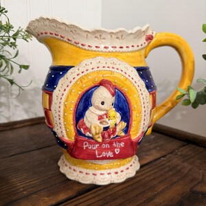 May include: A decorative ceramic pitcher with a yellow body, blue and red accents, and a white lace-trimmed rim. The pitcher features a central design of a chick and a baby chick, with the text "Pour on the Love".