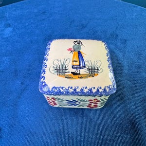 May include: A square ceramic box with a cream-colored lid featuring a hand-painted illustration of a person in a blue and yellow outfit standing in a field. The sides of the box are decorated with a repeating pattern of red flowers and blue leaves.