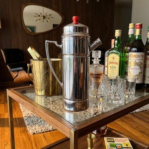 May include: A tall, polished chrome cocktail shaker with a red knob on the lid, positioned on a glass-topped bar cart. The shaker features a long handle and a spout, accompanied by a brass ice bucket, crystal glassware, and a cocktail recipe book.