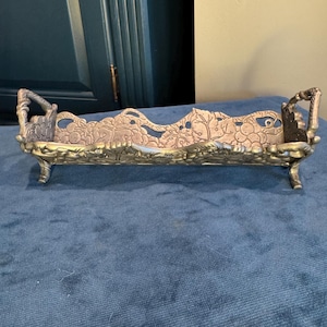 May include: An antique silver-toned metal tray with ornate floral and leaf detailing. The tray has a rectangular shape with curved handles on each end and sits on four small feet. The design features a wavy top edge.