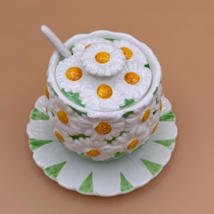 May include: A white ceramic sugar bowl with a matching lid and saucer, featuring a raised three-dimensional daisy pattern with yellow centers and green leaves. A small white ceramic spoon rests inside the bowl through a notch in the lid.