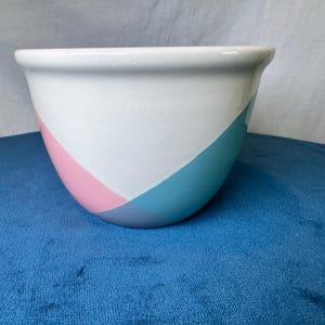 May include: A ceramic bowl with a smooth white upper section and a base featuring geometric color-blocked segments in pastel pink, light blue, and gray. The bowl has a wide, rounded opening and a curved body, presenting a clean, glossy finish.