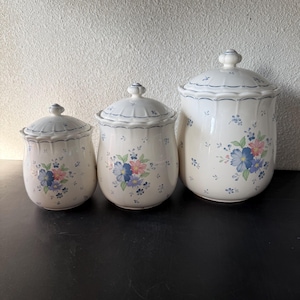 May include: Set of three ceramic canisters with lids. The canisters are white with a floral pattern of blue, pink, and purple flowers. Each canister has a scalloped edge and a small knob on the lid. The canisters are different sizes.