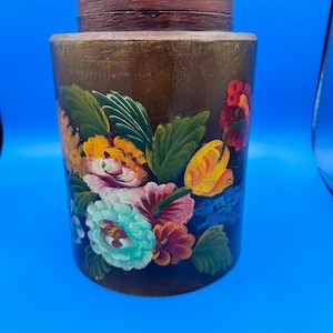 May include: A cylindrical wooden canister with a dark brown finish, featuring a hand-painted floral arrangement in shades of turquoise, yellow, red, and orange, accented by green leaves. The container includes a matching dark brown wooden lid.