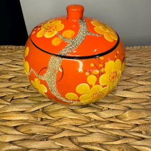 May include: A round, orange decorative jar with a lid. The jar is adorned with yellow floral designs and gold glitter accents. A black line encircles the base of the lid. The jar sits on a woven surface.