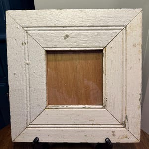 May include: Square, distressed white wooden frame with a natural wood center. The frame has a layered design with visible cracks in the paint, giving it a rustic appearance. The frame is supported by a black stand.