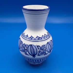 May include: A white ceramic vase with a flared rim and a rounded body, featuring hand-painted blue floral and geometric patterns. The design includes horizontal bands of scalloped lines and stylized fruit motifs encircling the center.