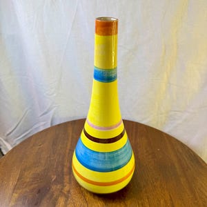 May include: A tall, slender ceramic vase with a bright yellow base color. The surface is decorated with horizontal stripes in shades of orange, light blue, dark brown, and soft pink, creating a vibrant, multi-colored pattern around the body.