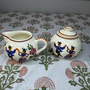 May include: Cream-colored ceramic creamer and sugar bowl set. The set features a folk art design of dancing figures in traditional attire. The creamer has a handle and spout, while the sugar bowl has a lid. Brown trim accents the edges.