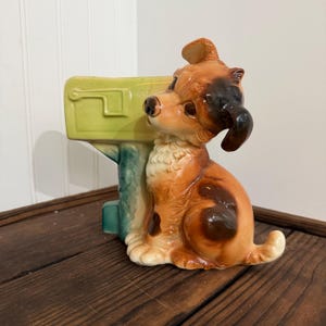 May include: A ceramic dog figurine with brown, black, and white fur sits next to a light green mailbox. The dog has a glossy finish and is posed in a seated position. The mailbox has a rectangular shape with a slot and a blue-green post.