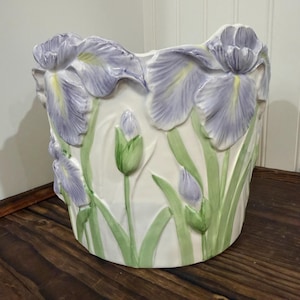 May include: A white ceramic planter featuring a raised, three-dimensional relief design of light purple iris flowers with yellow centers and green stems and leaves. The cylindrical vessel has a smooth, glossy finish and an uneven, scalloped top edge.