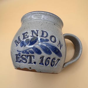 May include: A speckled light gray ceramic mug with a rounded body and a side handle. The front features blue embossed text reading MENDON and EST. 1667, with a blue painted leaf design positioned between the two lines of text on the mug surface.