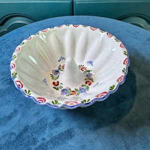 May include: A white ceramic fluted ring mold or bundt pan featuring a central cone and a hand-painted floral pattern in red, blue, and green around the base and along the scalloped rim, with a glossy finish and vertical ridges along the interior.