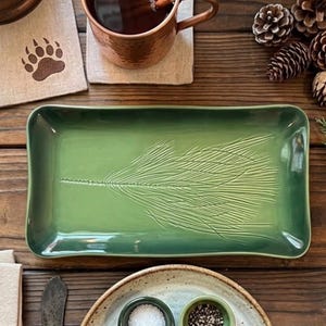 May include: A rectangular green ceramic serving tray featuring an embossed pine needle branch design. The tray has a smooth, glossy finish with slightly raised edges and a deep forest green color, centered on a rustic wooden surface.