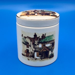 May include: A cylindrical ceramic canister with a matching lid, featuring a gold-trimmed rim. The exterior displays a detailed illustration of a multi-story building with green roofs and the text Four Ways Inn, set against a cream-colored background.