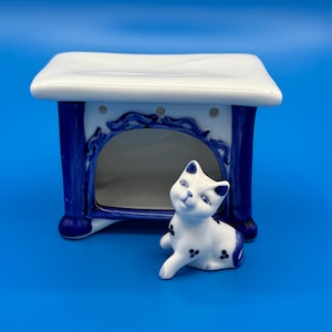 May include: A white and blue ceramic oil burner featuring a rectangular top and a hollow arched base, accompanied by a small white and blue ceramic cat figurine with painted spots, positioned in front of the burner on a solid blue surface.