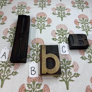 May include: Four wooden letterpress printing blocks, including the letters B, b, and C, are displayed on a floral patterned fabric. The blocks are in various sizes and shades of brown, with the letter "b" having a contrasting gold-colored interior.