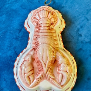 May include: A ceramic lobster-shaped mold featuring a cream-colored base with hand-painted reddish-orange accents along the edges and the lobster relief. The mold includes a small metal ring at the top for hanging, showcasing a detailed crustacean.