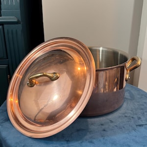 May include: A copper-clad cooking pot with a lid, set on a blue surface. The pot has a shiny copper exterior and a stainless steel interior. The lid and handles are a gold color.