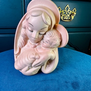 May include: A vintage pink ceramic figurine depicting a mother and child. The mother figure wears a halo and cradles the baby. The figurine has a glossy finish and is set against a blue background.