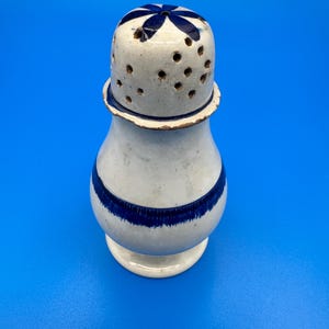 May include: A ceramic salt or pepper shaker with a cream-colored glaze and a rounded body. It features a dark blue horizontal band around the center and a matching blue flower design on the top lid, which is perforated with multiple small holes.