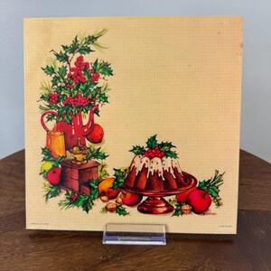 May include: A vintage square print featuring a festive holiday scene. A red teapot and a cake are surrounded by apples, nuts, and holly. The print has a cream-colored background and is displayed on a wooden surface.