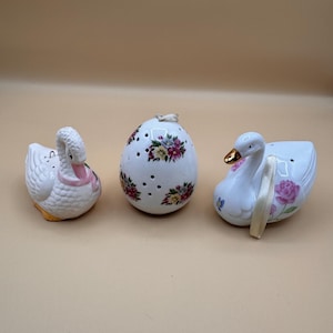 May include: A set of three ceramic items including a white swan figurine with a pink neck accent, a white egg-shaped ornament with floral patterns and small ventilation holes, and a white swan figurine featuring a gold beak and pink rose details.