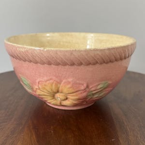 May include: A vintage pink ceramic bowl with a floral design. The bowl features a raised floral pattern in the center, with a rope-like design around the rim. The interior is a cream color.