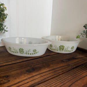 May include: Two oval, white casserole dishes with green floral designs. The dishes have a simple, elegant shape with handles on each side. The green floral pattern is a repeating design of flowers and leaves.